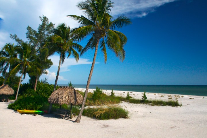 Tropical beach with palm trees, straw huts, and a kayak on white sand under a clear blue sky.