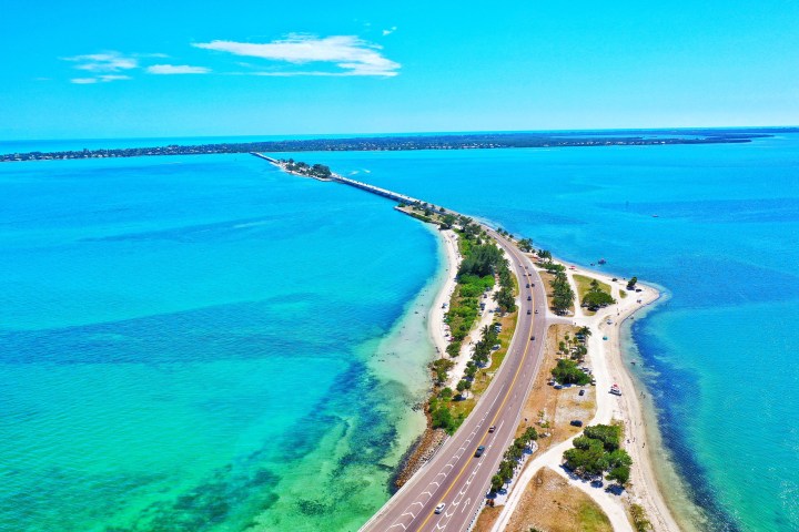 Aerial view of a road stretching through a turquoise sea, lined with trees, connecting two land areas.